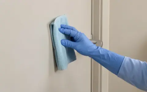Gloved hand wiping the door surface beside a lever handle with a microfiber cloth